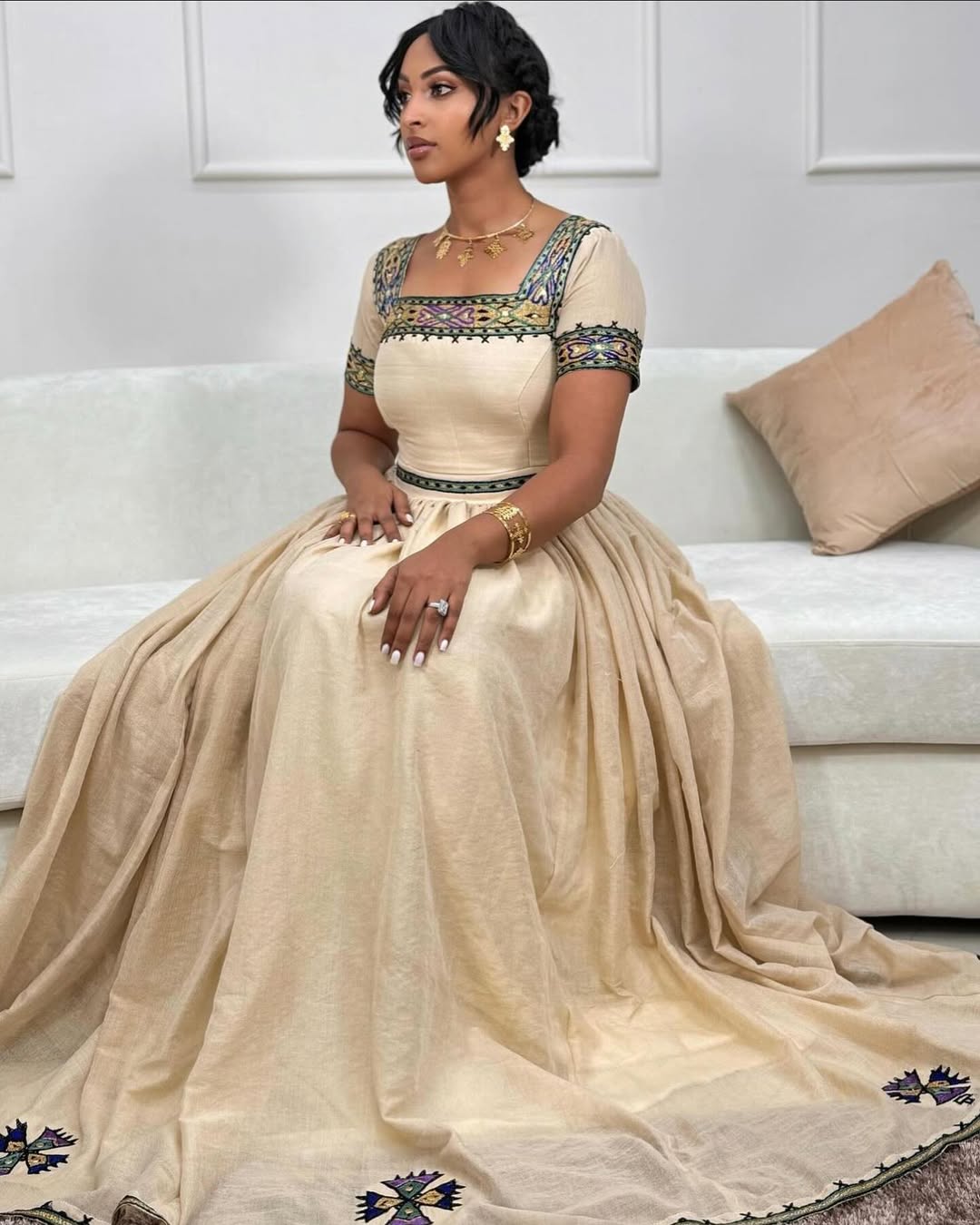 Woman in a beige saree with intricate designs sitting on a couch. New Beige Zuria Habesha Kemis Modern Handcrafted Ethiopian Dress ethgebya gebeya