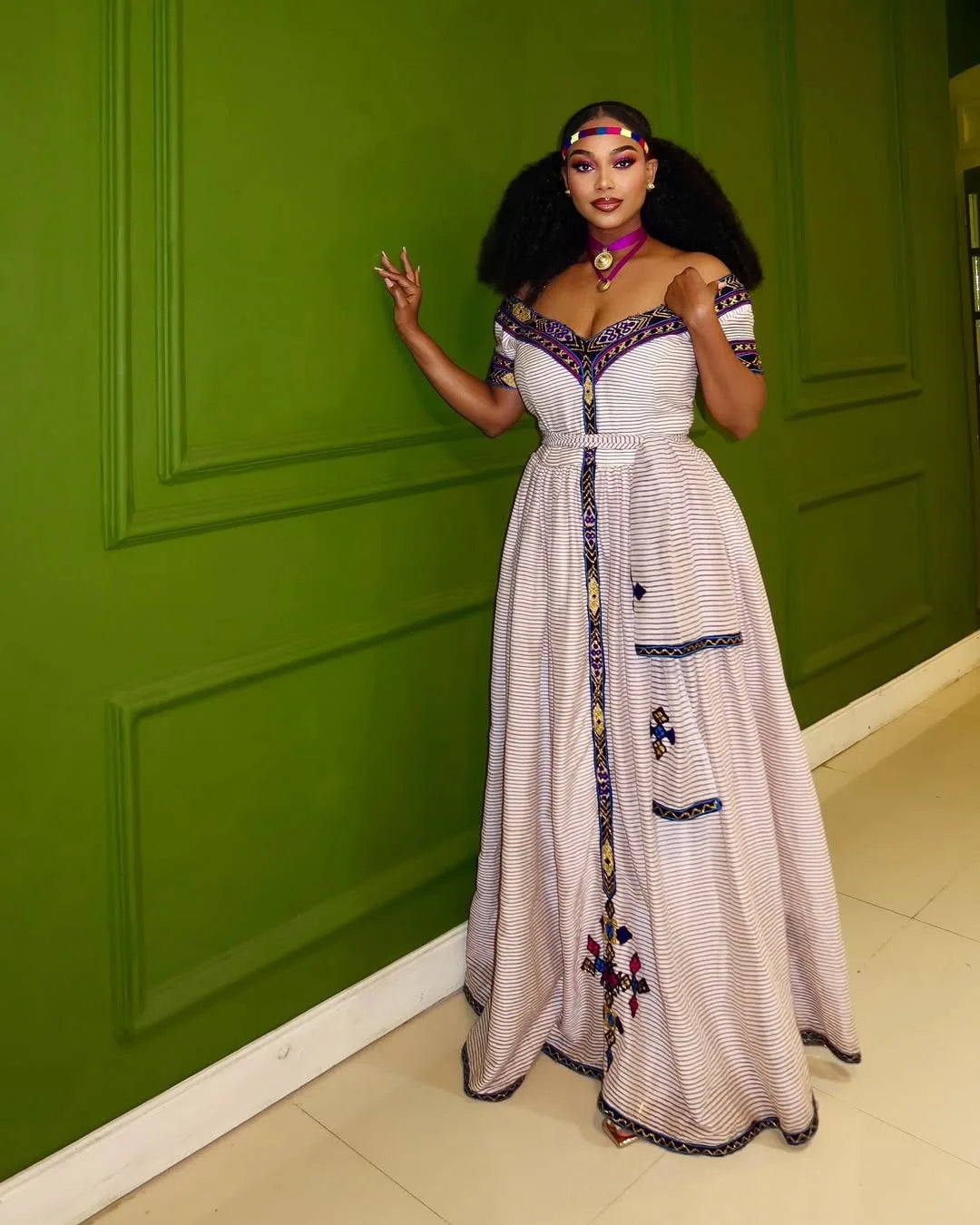 Woman in a white dress with embroidery standing against a green wall. New Striped Zuria Kemis | Modern Handcrafted Ethiopian Dress