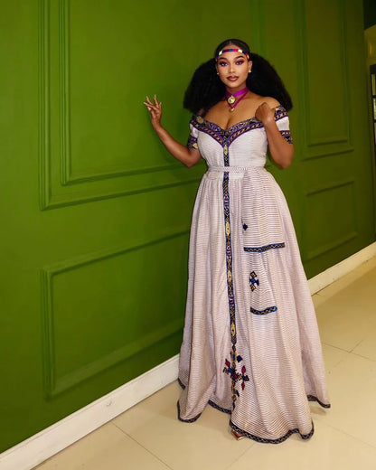 Woman in a white dress with embroidery standing against a green wall. New Striped Zuria Kemis | Modern Handcrafted Ethiopian Dress