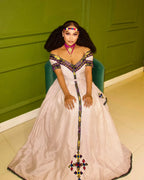 Woman in a white dress with colorful embellishments sitting on a green chair against a green wall. New Striped Zuria Kemis | Modern Handcrafted Ethiopian Dress