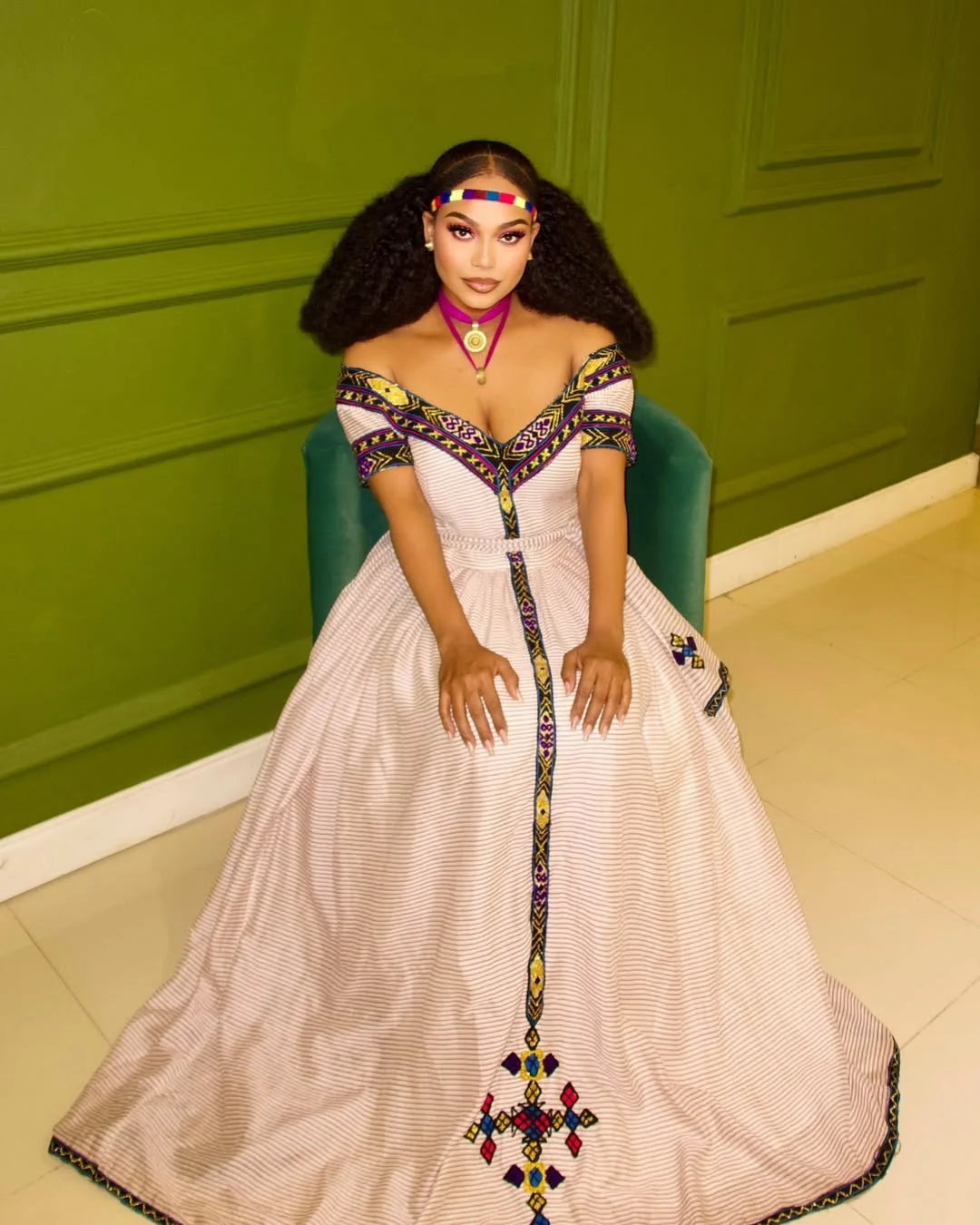 Woman in a white dress with colorful embellishments sitting on a green chair against a green wall. New Striped Zuria Kemis | Modern Handcrafted Ethiopian Dress