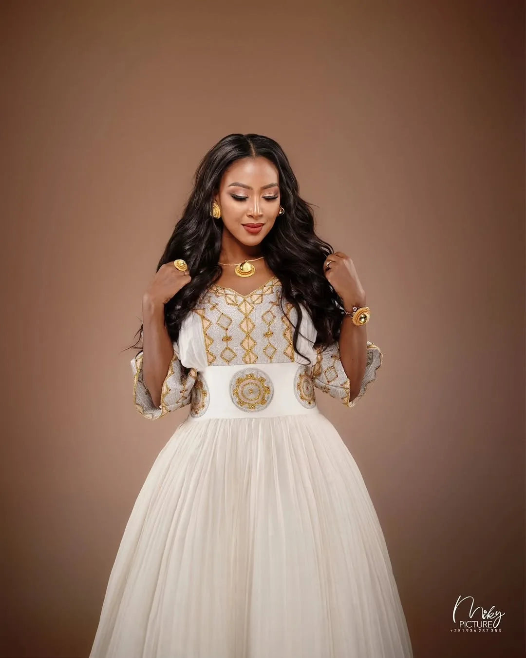 Woman in a white and gold dress with jewelry against a brown background. Golden Modern Zuria Habesha Kemis Elegant Ethiopian Traditional Dress ethgebya