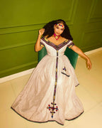 Woman in a white dress with colorful embroidery sitting on a green chair against a green wall. New Striped Zuria Kemis | Modern Handcrafted Ethiopian Dress