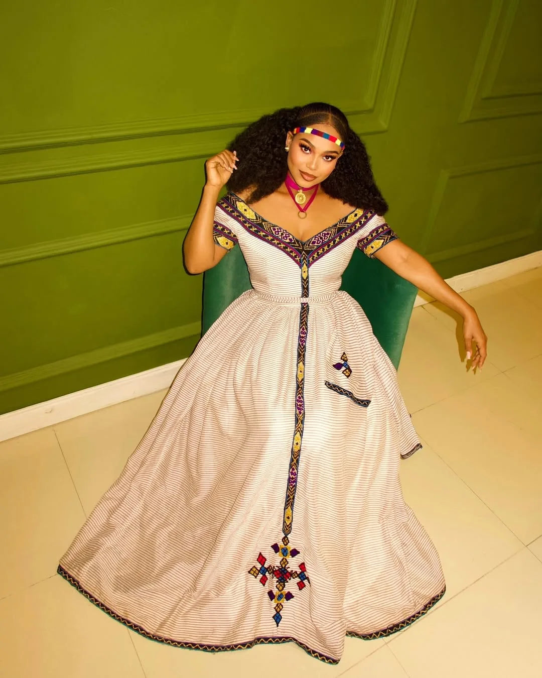 Woman in a white dress with colorful embroidery sitting on a green chair against a green wall. New Striped Zuria Kemis | Modern Handcrafted Ethiopian Dress