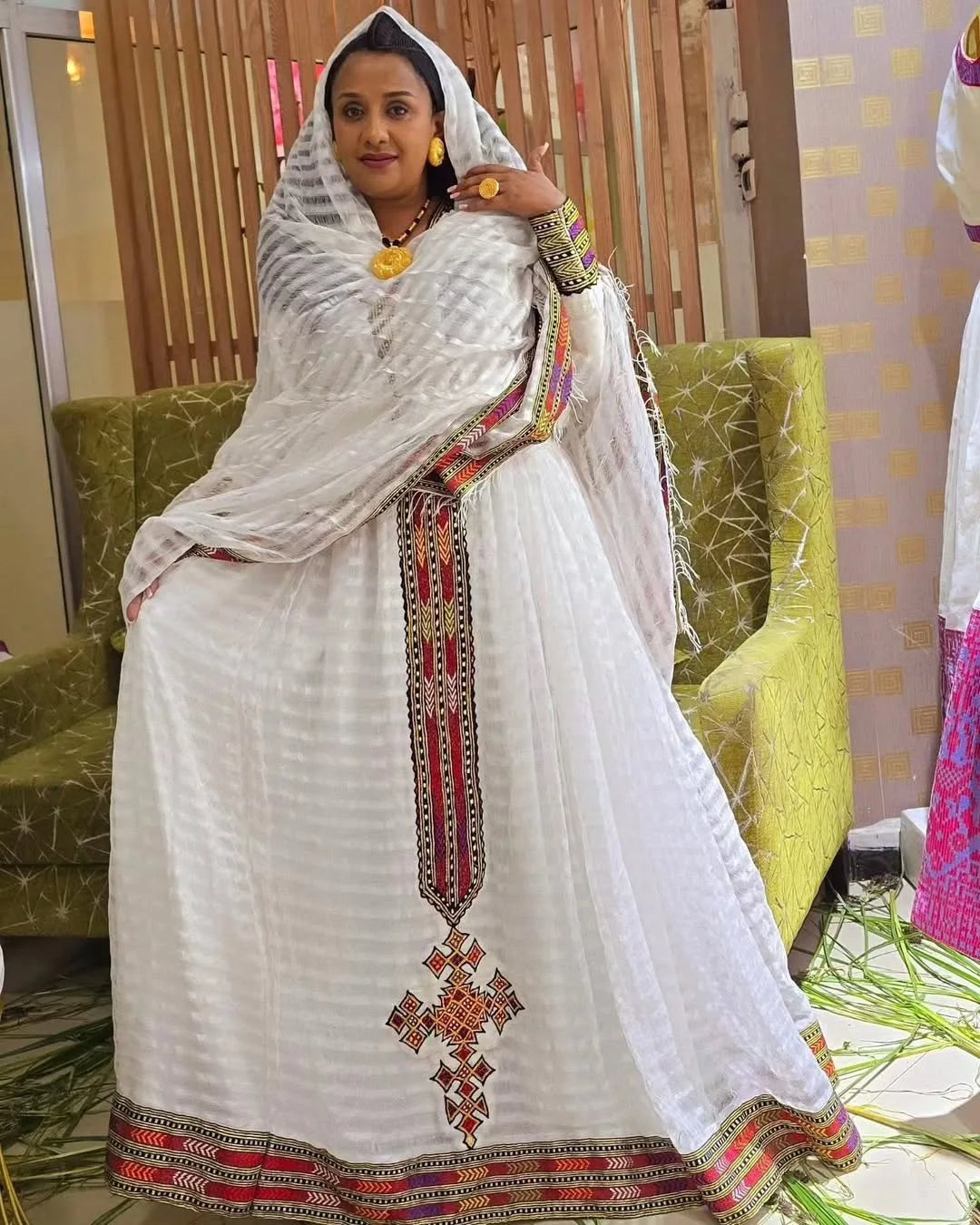 Woman in a white traditional outfit with colorful patterns sitting on a green couch. Elegant Zuria Habesha Kemis New Modern Ethiopian Traditional Dress ethgebya gebeya