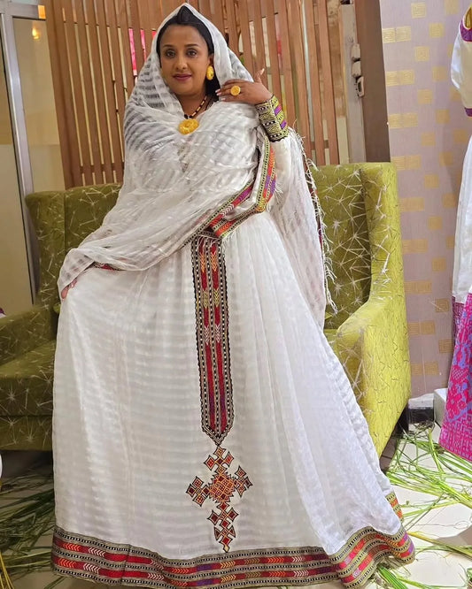 Woman in a white traditional outfit with colorful patterns sitting on a green couch. Elegant Zuria Habesha Kemis New Modern Ethiopian Traditional Dress ethgebya gebeya