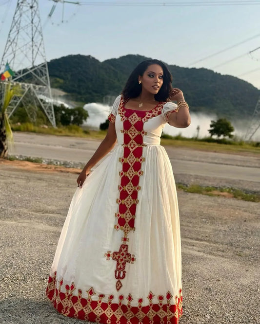 Woman wearing a white dress with red patterns standing on a road with mountains in the background.