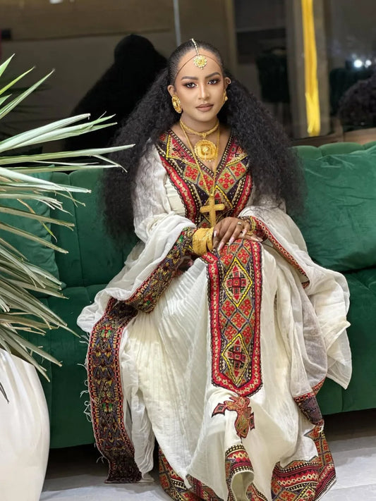 Woman in traditional embroidered outfit sitting on a green couch. axum habesha kemis Shop Now & Enjoy Free Worldwide Shipping – Fast Delivery to USA, Canada, UK, and Europe!