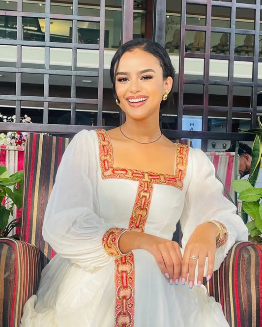 Woman in a white dress with red and gold embroidery sitting on a striped chair.