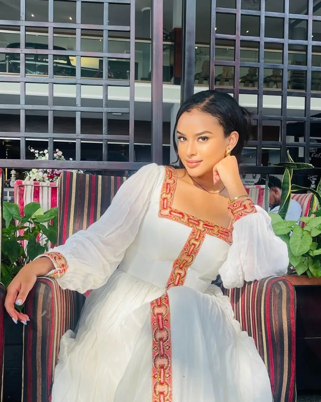 Woman in a white dress with red and gold embroidery sitting on a colorful chair.