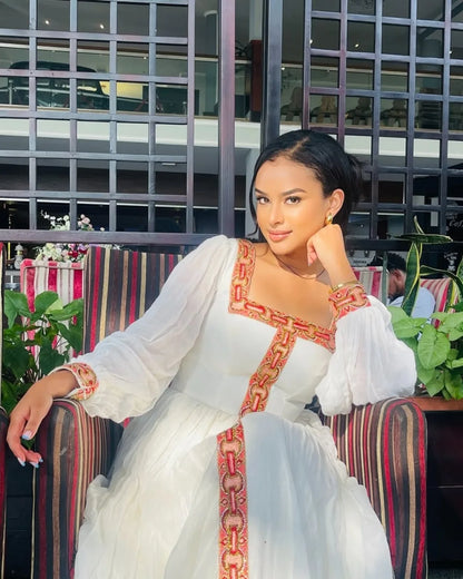 Woman in a white dress with red and gold embroidery sitting on a colorful chair.