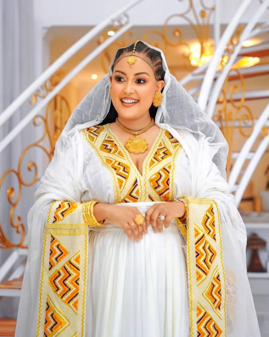 Woman in traditional white and gold outfit with decorative patterns, standing indoors. Orange & Yellow Zuria Habesha Kemis Modern Ethiopian Traditional Dress Ethgebya