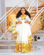 Woman in a white and gold traditional outfit standing in a decorative interior setting. Orange & Yellow Zuria Habesha Kemis Modern Ethiopian Traditional Dress Ethgebya