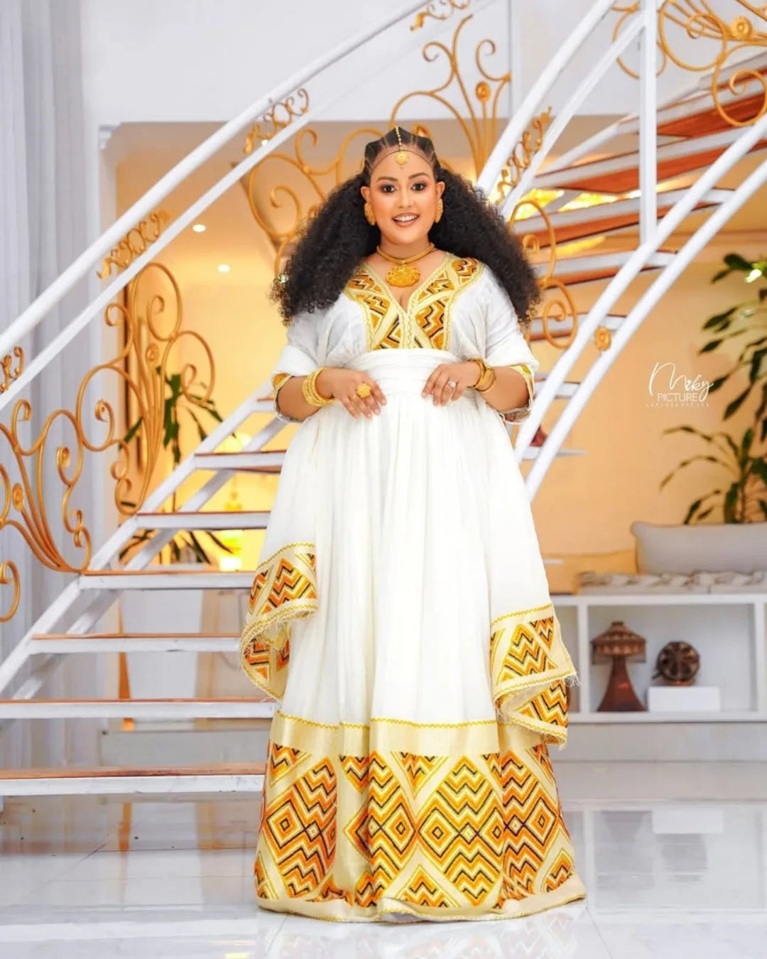 Woman in a white and gold traditional outfit standing in a decorative interior setting. Orange & Yellow Zuria Habesha Kemis Modern Ethiopian Traditional Dress Ethgebya