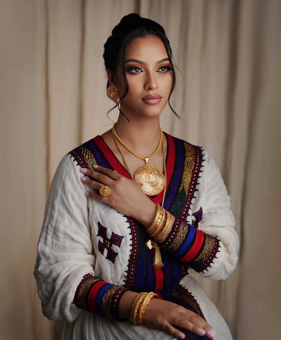 Woman wearing traditional attire with jewelry against a beige curtain background. Elegant Ethiopian & Eritrean Axum Habesha Dress Ethgebya