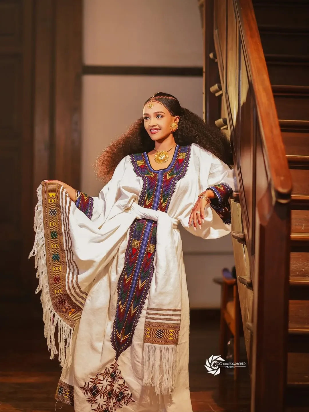 Woman in a traditional embroidered dress standing in a room with wooden stairs. New Ethiopian & Eritrean Axum Habesha Dress Handwoven Tilet Kemis Ethiopia Gebeya Ethgebya