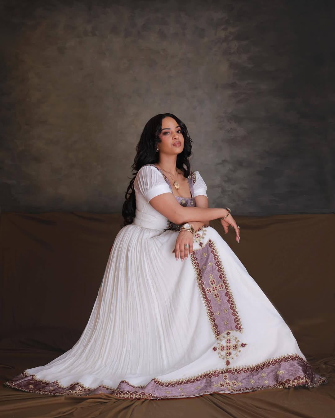 Woman in a white and purple traditional outfit sitting on a brown couch against a textured brown wall. Rose Gold Zuria Habesha Kemis Handcrafted Modern Ethiopian Dress Ethiopia Gebeya Ethgebya