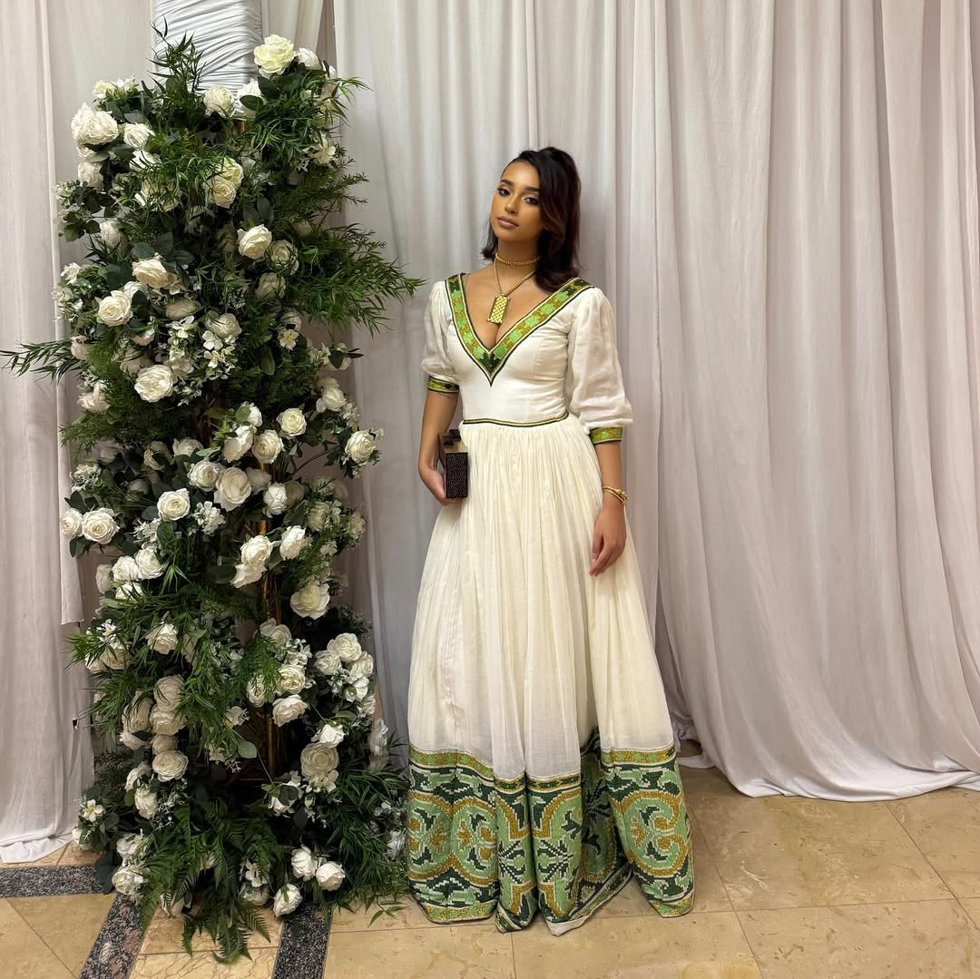 Woman in a white and green dress standing next to a floral arrangement against a white curtain. Elegant Green Zuria Habesha Kemis Modern Ethiopian Traditional Dress thgebya gebeya