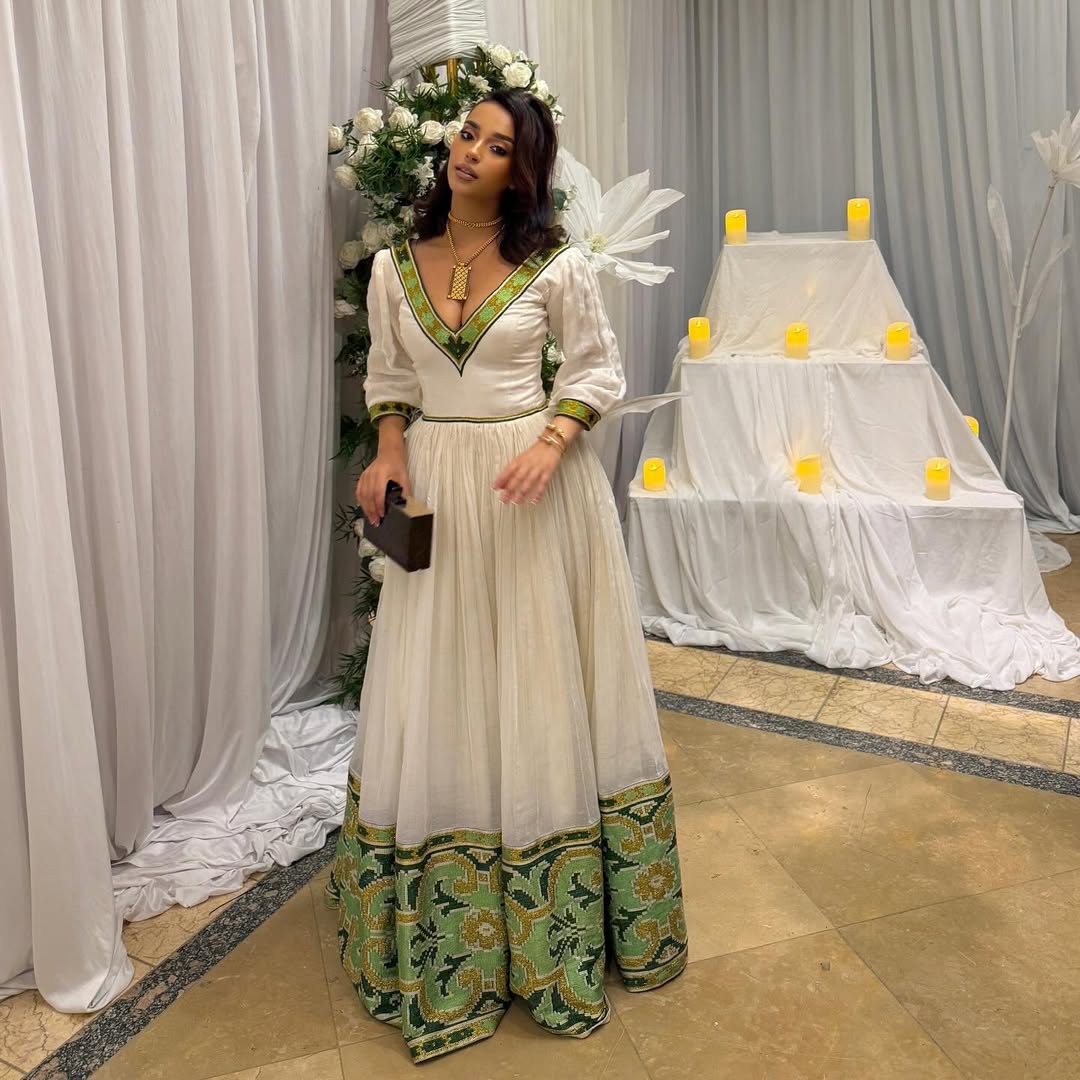 Woman in a white and green dress standing in front of a decorated setup with flowers and candles. Elegant Green Zuria Habesha Kemis Modern Ethiopian Traditional Dress thgebya gebeya