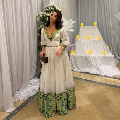 Woman in a white and green dress standing in front of a decorated setup with flowers and candles. Elegant Green Zuria Habesha Kemis Modern Ethiopian Traditional Dress thgebya gebeya