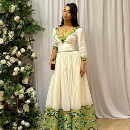 Woman in a white and green dress standing next to floral decorations against a white curtain. Elegant Green Zuria Habesha Kemis Modern Ethiopian Traditional Dress thgebya gebeya