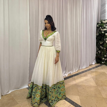 Woman in a white and green dress standing against a white curtain backdrop. Elegant Green Zuria Habesha Kemis Modern Ethiopian Traditional Dress thgebya gebeya