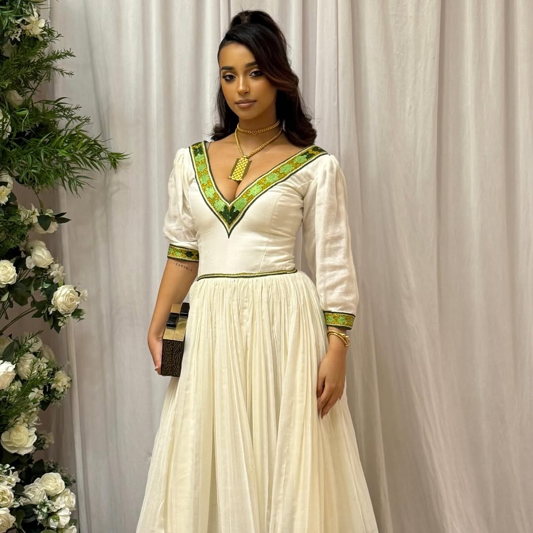Woman in a white dress with green accents standing next to floral decorations against a plain background Elegant Green Zuria Habesha Kemis Modern Ethiopian Traditional Dress thgebya gebeya