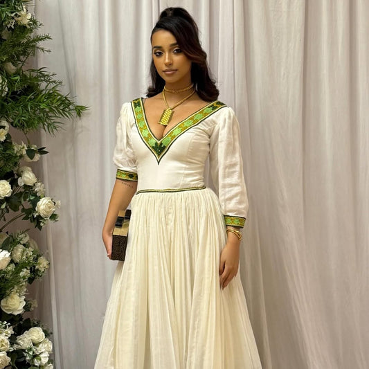 Woman in a white dress with green accents standing next to floral decorations against a plain background Elegant Green Zuria Habesha Kemis Modern Ethiopian Traditional Dress thgebya gebeya