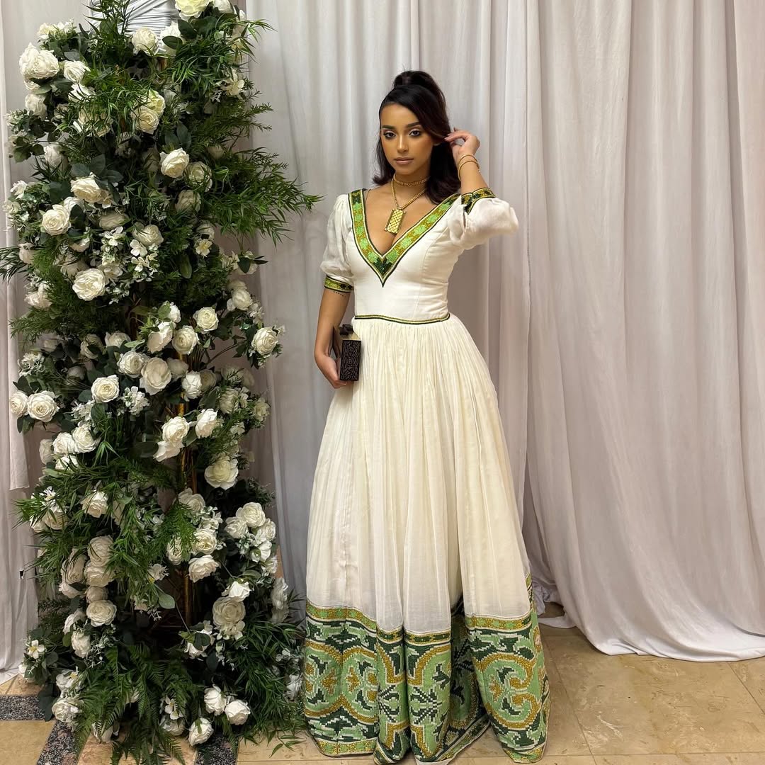 Woman in a white and green dress standing next to a floral arrangement against a white curtain. Elegant Green Zuria Habesha Kemis Modern Ethiopian Traditional Dress thgebya gebeya