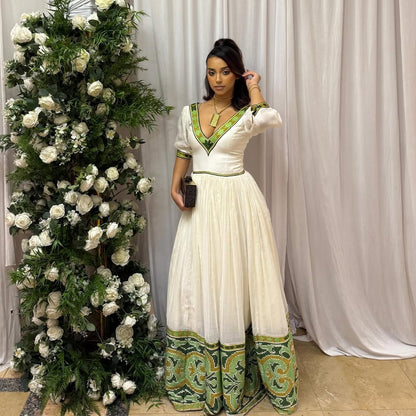 Woman in a white and green dress standing next to a floral arrangement against a white curtain. Elegant Green Zuria Habesha Kemis Modern Ethiopian Traditional Dress thgebya gebeya