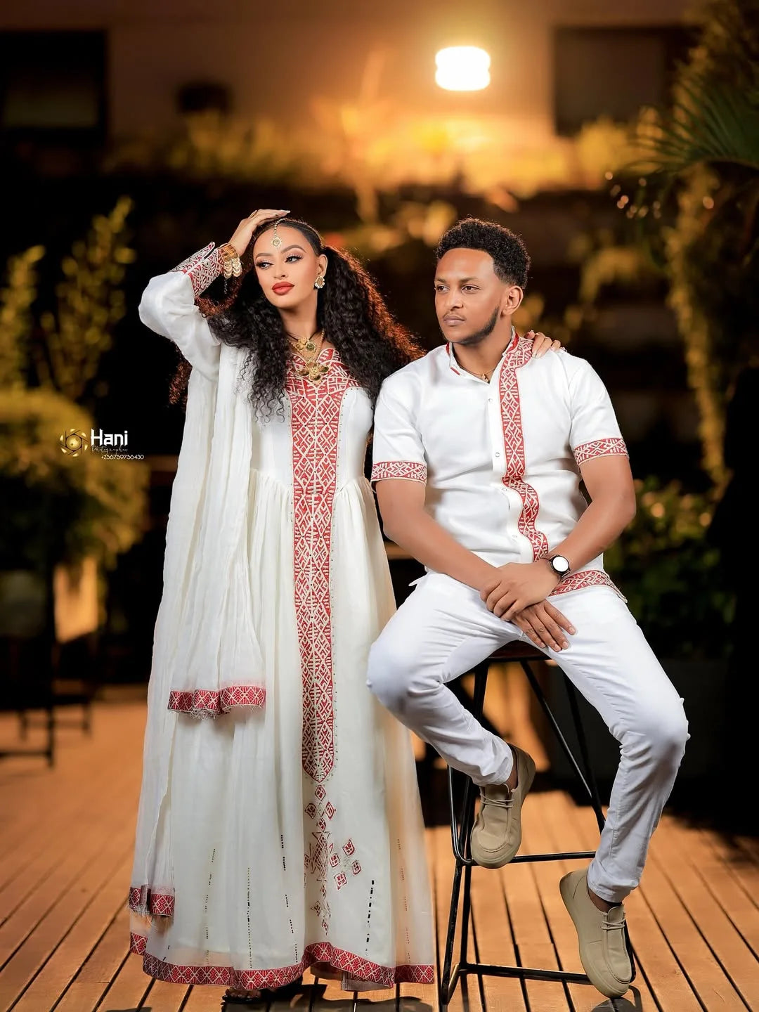 Two people in formal attire sitting together on a wooden floor with plants in the background. Red Ethiopian Couple Set ethgebya