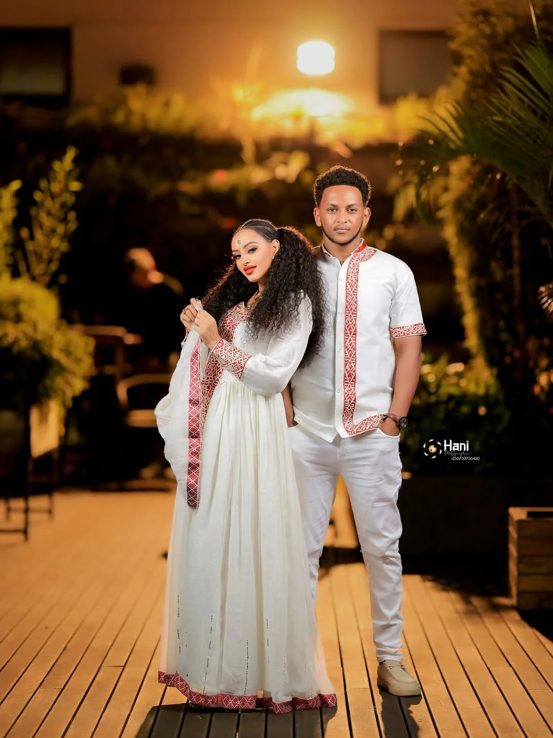 Couple in formal attire standing on a wooden deck with plants and warm lighting in the background. Red Ethiopian Couple Set ethgebya