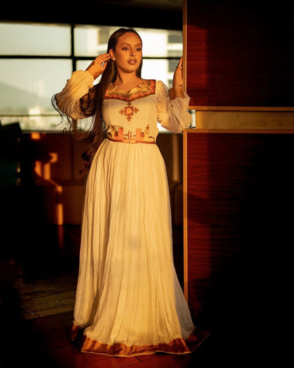Woman in a long, light-colored dress with intricate patterns standing in a softly lit room. Orange & Golden Zuria Kemis | Modern Handcrafted Ethiopian Dress