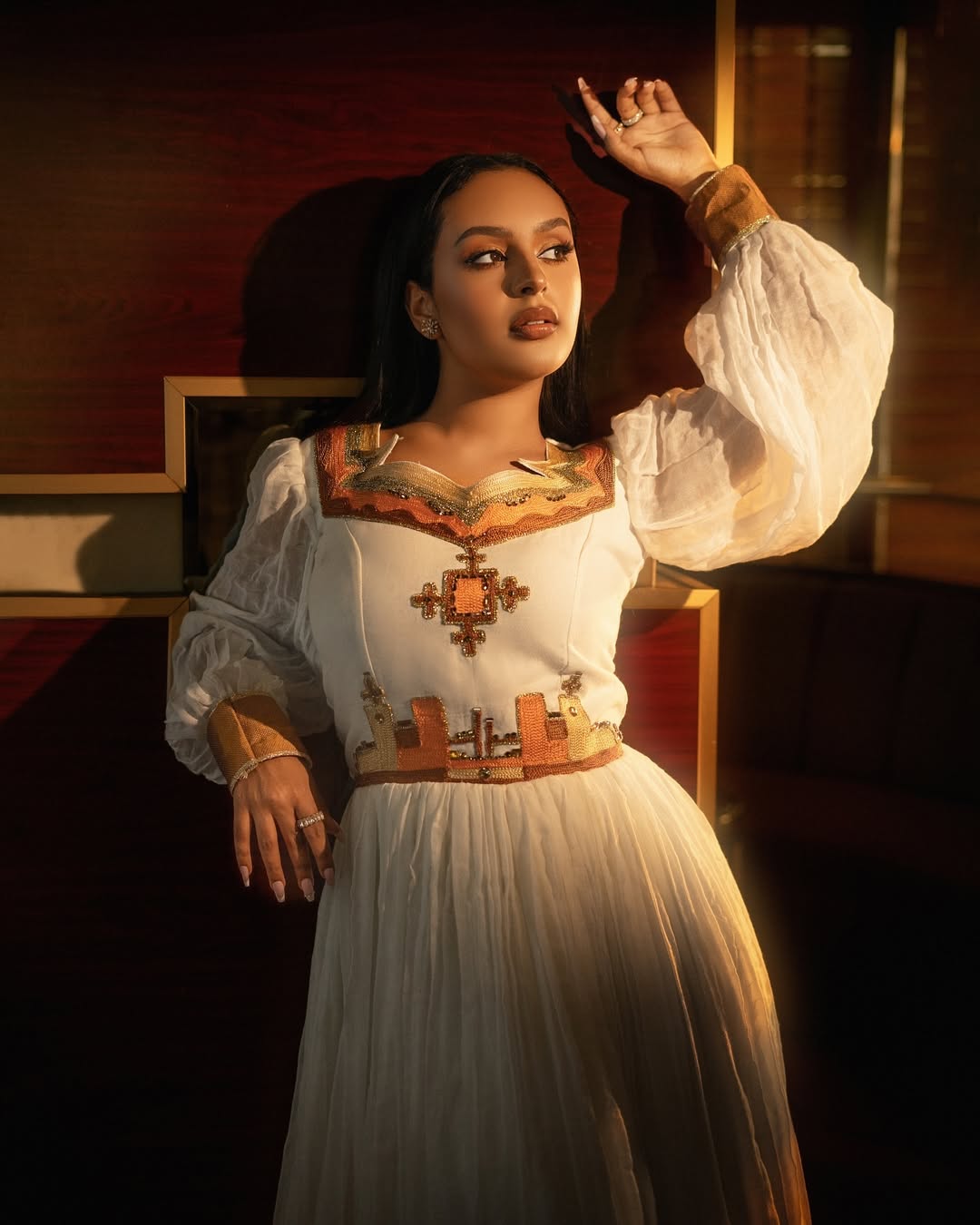 Woman in a white dress with intricate patterns in a dimly lit room Orange & Golden Zuria Kemis | Modern Handcrafted Ethiopian Dress