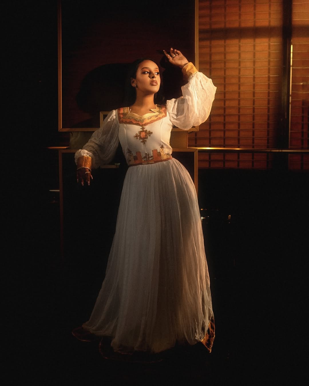 Woman in a traditional outfit with intricate patterns in a dimly lit room. Orange & Golden Zuria Kemis | Modern Handcrafted Ethiopian Dress