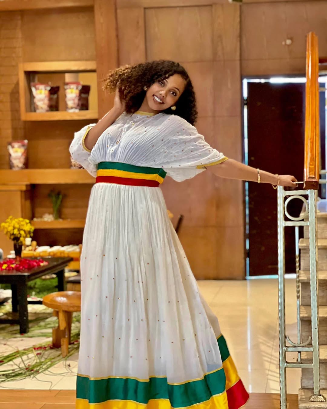Woman in a white dress with colorful hem in a room setting Green, Yellow & Red Habesha Zuria Kemis Modern Ethiopian Dress ethgebya gebeya