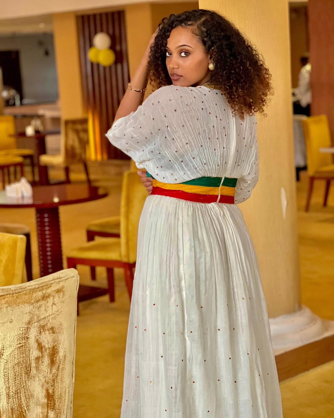 Woman in a white dress with a red belt standing in a restaurant. Green, Yellow & Red Habesha Zuria Kemis Modern Ethiopian Dress ethgebya gebeya