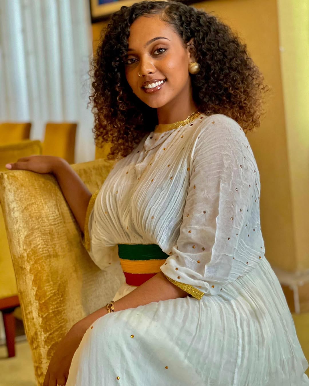 Woman in a white traditional outfit sitting on a yellow couch indoors. Green, Yellow & Red Habesha Zuria Kemis Modern Ethiopian Dress ethgebya gebeya