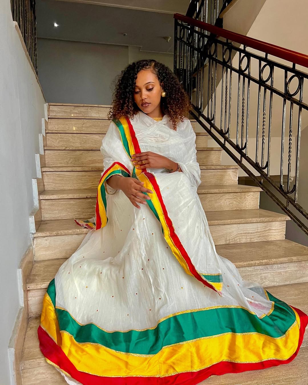 Woman in a white traditional outfit with colorful borders sitting on stairs. Green, Yellow & Red Habesha Zuria Kemis Modern Ethiopian Dress ethgebya gebeya