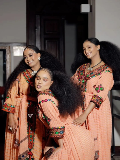 Three women in peach dresses with intricate patterns posing together indoors. Gold Sand Ethiopian & Eritrean Axum Habesha Dress Ethgebya