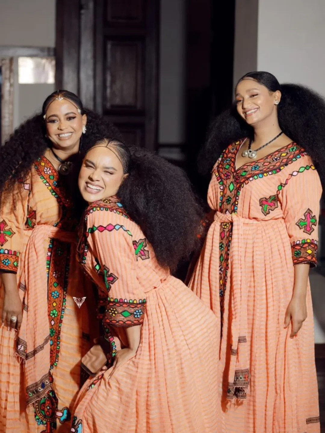Three women in traditional peach dresses with intricate patterns posing together. Gold Sand Ethiopian & Eritrean Axum Habesha Dress Ethgebya