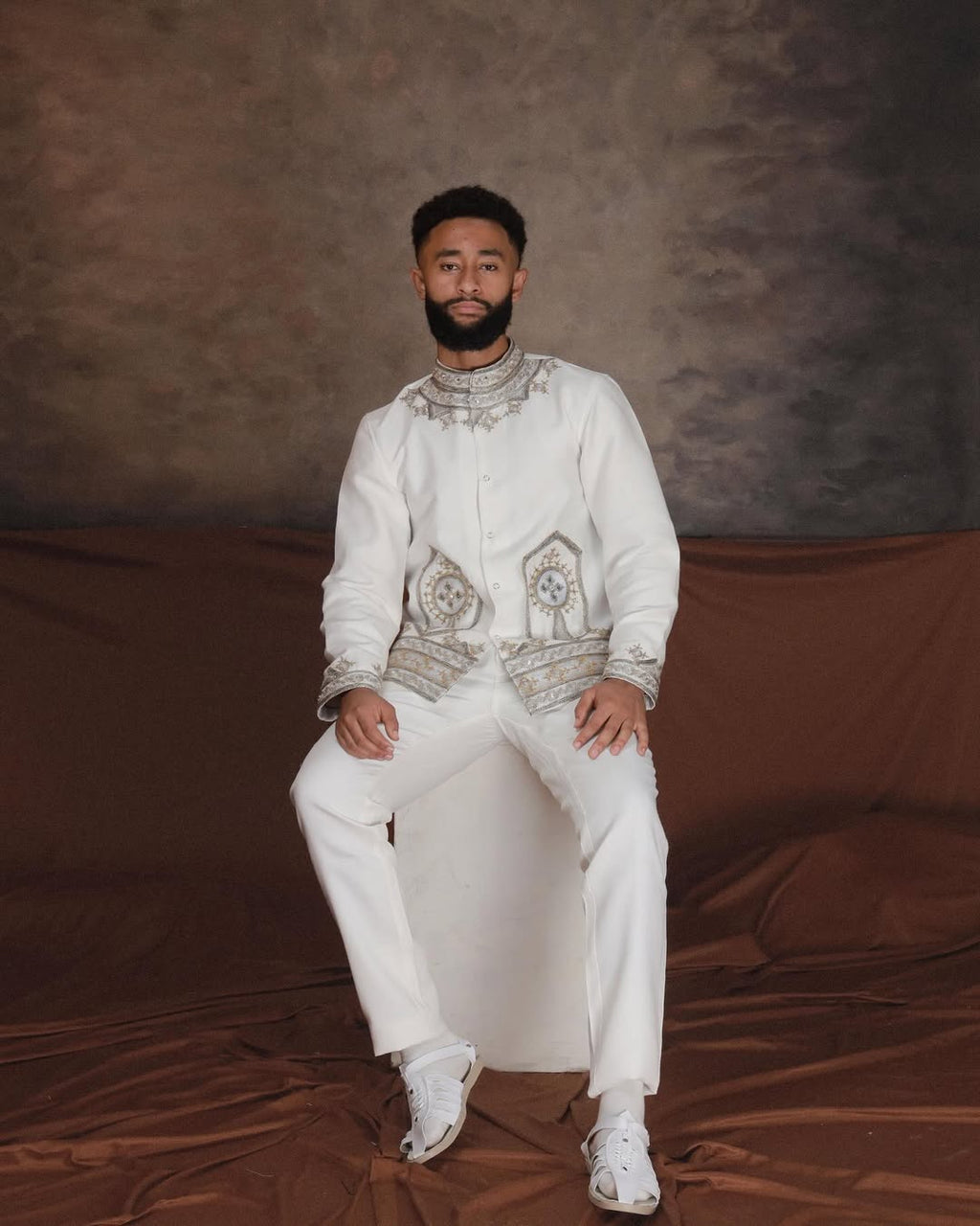 Man wearing a traditional outfit with intricate patterns sitting on a brown draped background. Ethiopian Men’s Traditional Outfit New Custom Handwoven Shirt ethgebya gebeya
