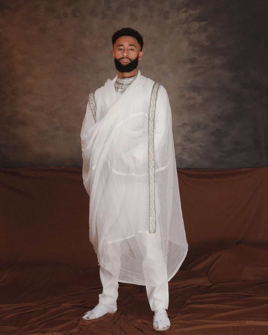 Man wearing a white traditional outfit against a brown backdrop. Ethiopian Men’s Traditional Outfit New Custom Handwoven Shirt ethgebya gebeya