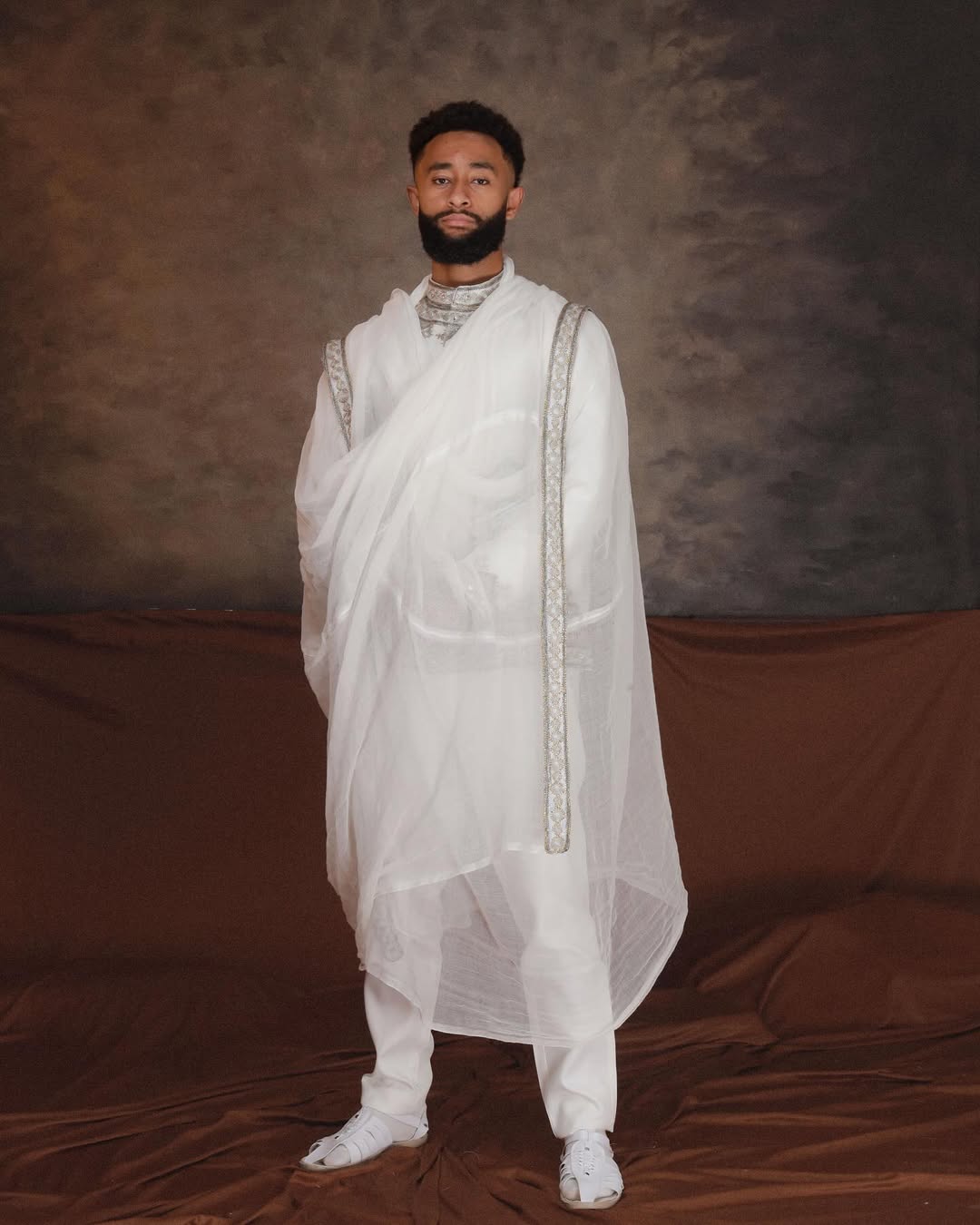 Man wearing a white traditional outfit against a brown backdrop. Ethiopian Men’s Traditional Outfit New Custom Handwoven Shirt ethgebya gebeya