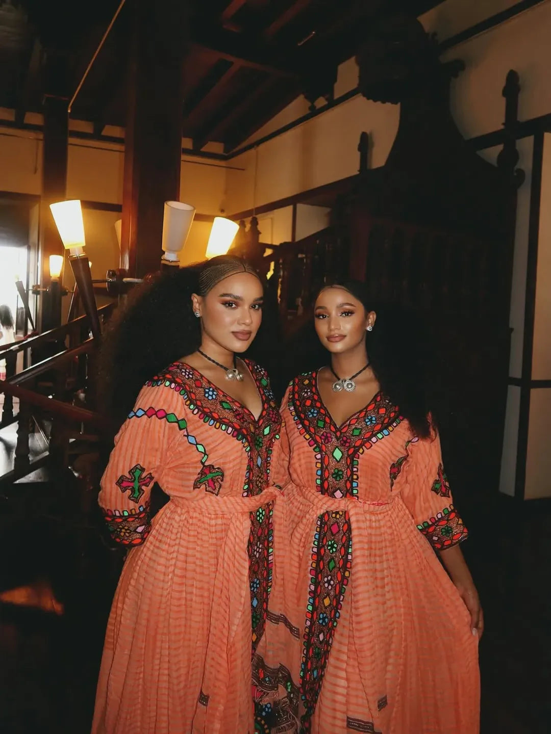 Two women in traditional orange dresses with intricate patterns standing in a dimly lit room. Gold Sand Ethiopian & Eritrean Axum Habesha Dress Ethgebya