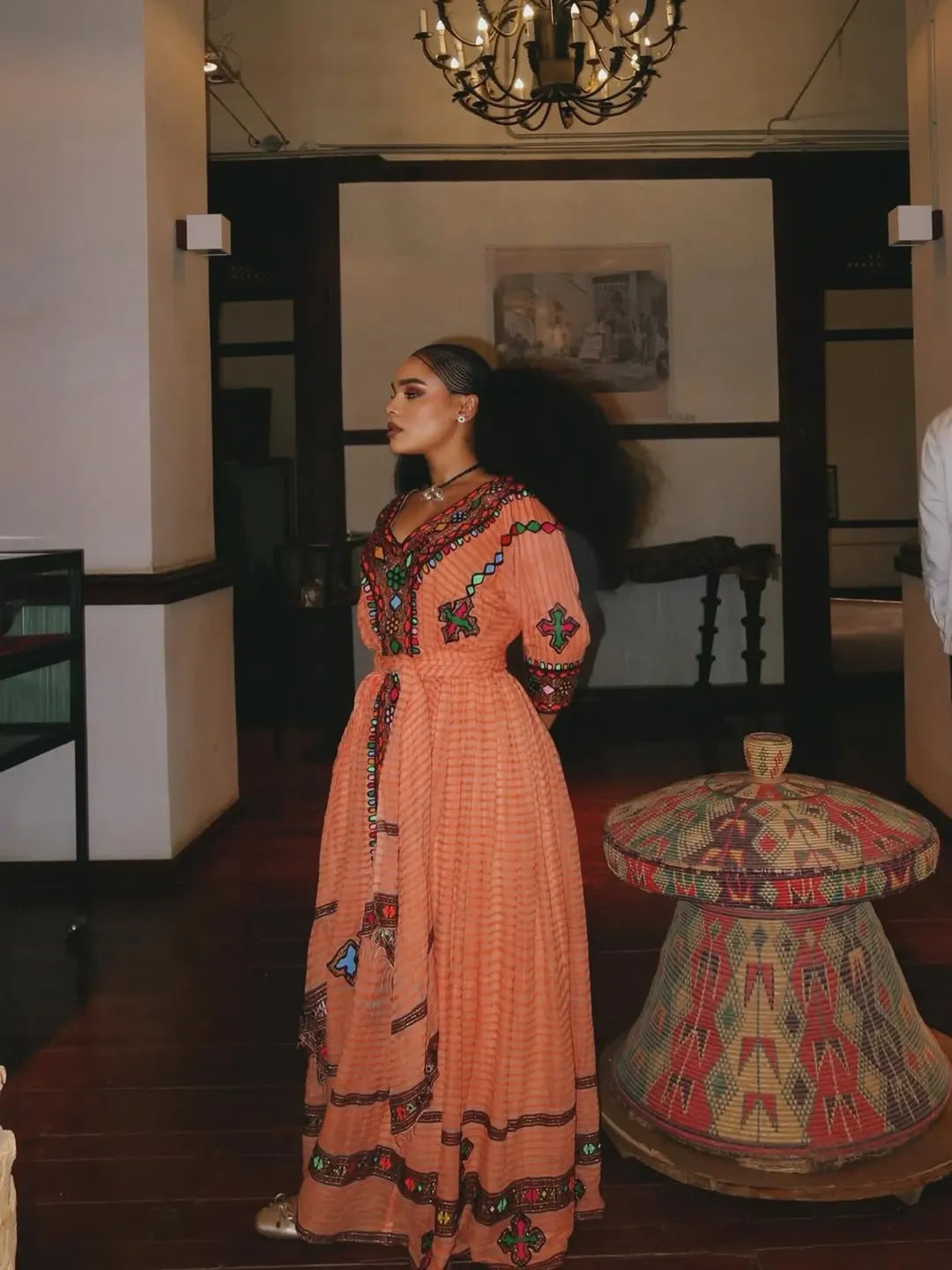 Woman in a long, patterned dress standing in an indoor setting with a chandelier and decorative table. Gold Sand Ethiopian & Eritrean Axum Habesha Dress Ethgebya