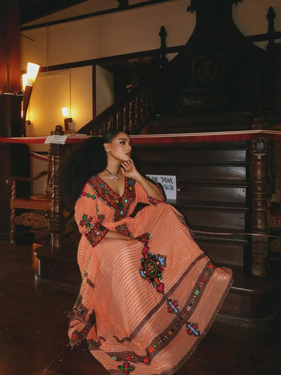Woman in a traditional outfit sitting on a staircase in a dimly lit room. Gold Sand Ethiopian & Eritrean Axum Habesha Dress Ethgebya