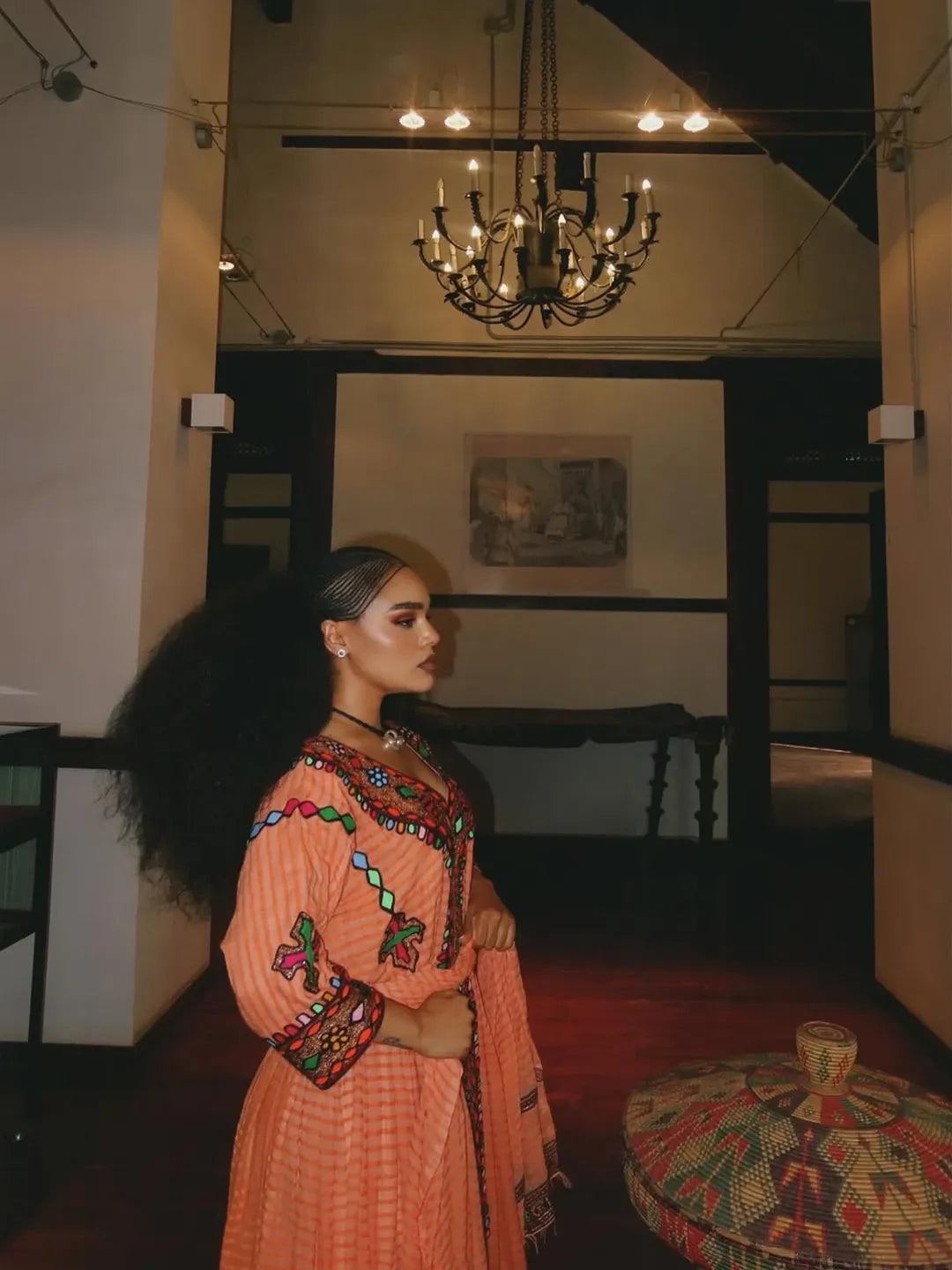 Woman in an ornate dress standing in a room with a chandelier and framed picture on the wall. Gold Sand Ethiopian & Eritrean Axum Habesha Dress Ethgebya