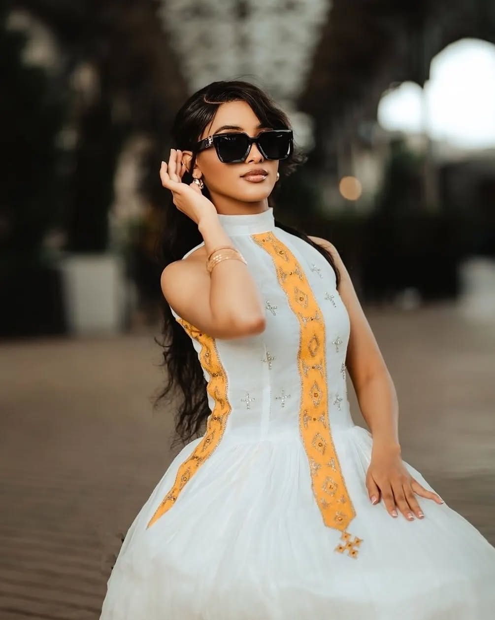 Woman wearing a white dress with orange accents and sunglasses outdoors. Yellow Modern Zuria Habesha Kemis Handcrafted Ethiopian Dress ethgebya gebeya usa habesha kemis