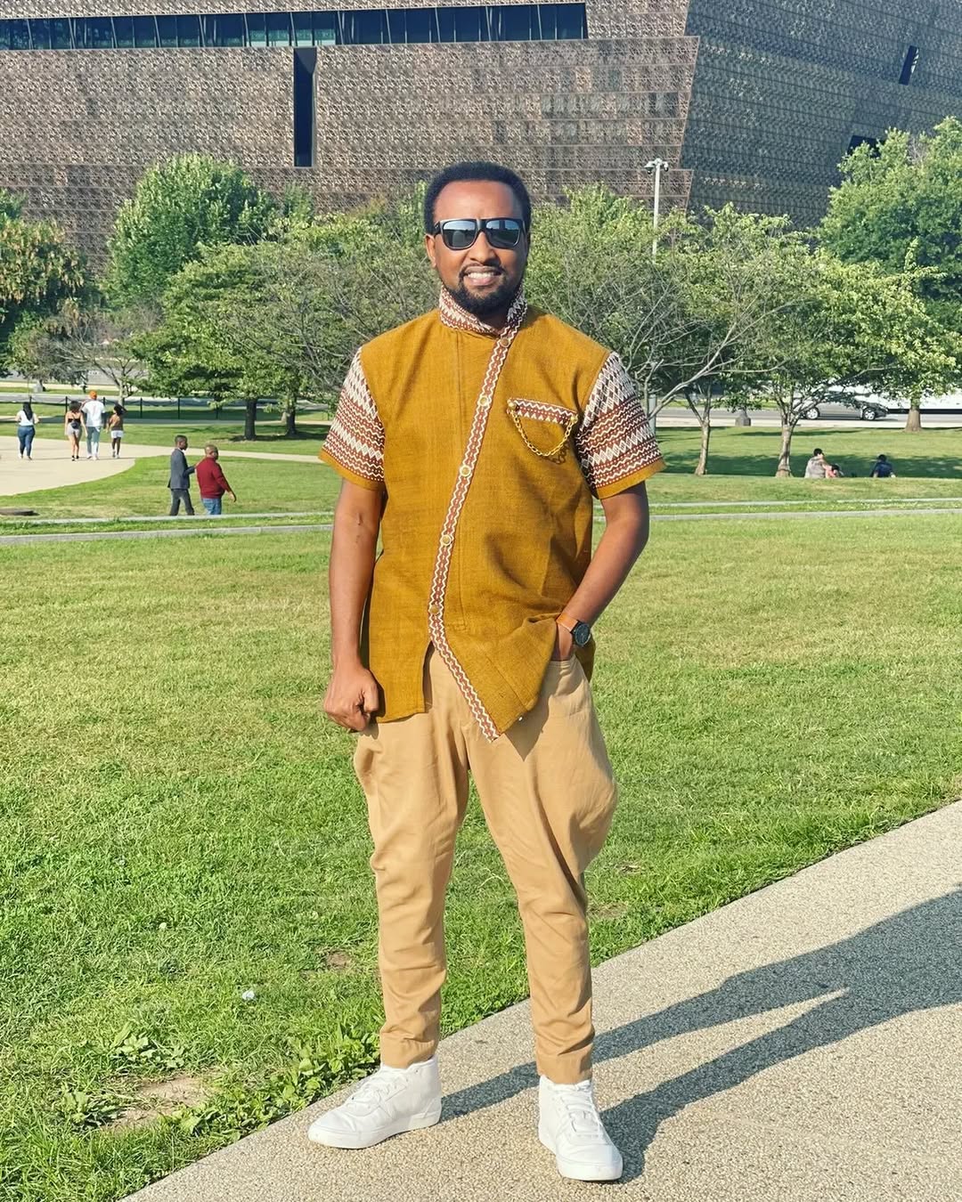Man in a mustard yellow shirt and beige pants standing on a sidewalk with a park in the background rown Modern Habesha Men’s Shirt Stylish Ethiopian Traditional Wear ethgebya gebeya usa habesha men